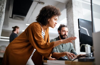 woman standing and pointing at computer screen that a man is sitting in front of
