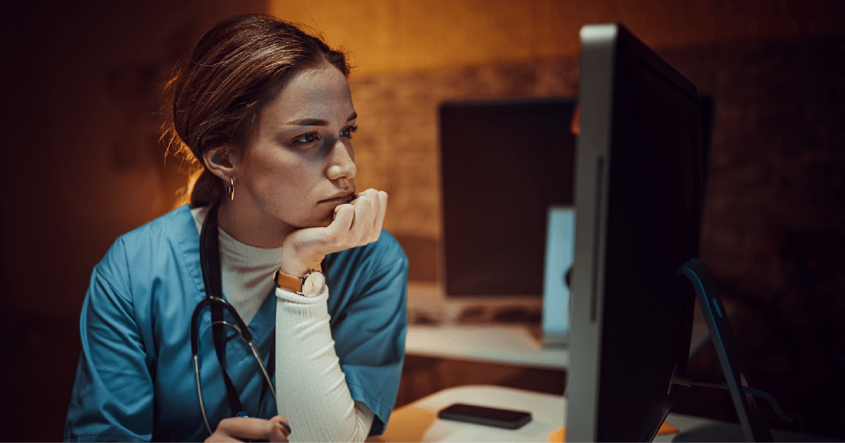 A female healthcare employee in scrubs looks concerned at her computer screen. 